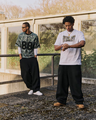To mænd poserer udendørs i urban omgivelser med oversize streetwear. Den ene bærer en camouflagemønstret Everlast trøje med tallet "88" og hvide sneakers, mens den anden har en hvid Everlast t-shirt med grønt logo og kigger på sit ur iført brune støvler. Baggrunden består af beton, glas og efterårsfarvede træer.