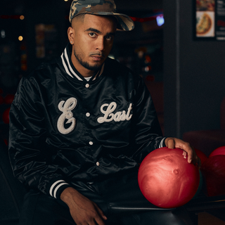 En mand sidder i en bowlinghal iført en sort satin varsity-jakke med teksten "E Last" i hvid skrift og en camouflagemønstret kasket. Han hviler hånden på en rød bowlingkugle og kigger direkte mod kameraet med et seriøst udtryk.