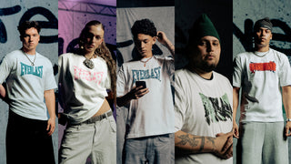 Fem personer står foran en grafittidækket bagvæg, alle iført hvide Everlast t-shirts med forskellige farvede logoer i blå, rød, grøn og sort. De poserer med individuelle udtryk og stilarter, hvilket skaber en varieret og ungdommelig streetwear-stemning.