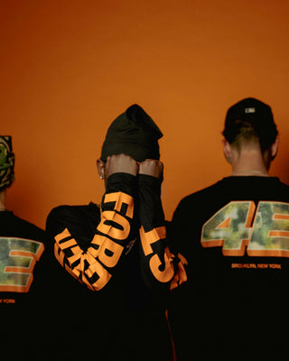 Tre mænd står foran en orange baggrund iført sorte Everlast-trøjer. De to yderste vender ryggen til og viser stort camouflageinspireret “4E” tryk med teksten “BROOKLYN, NEW YORK”, mens manden i midten har hænderne løftet og viser ærmeteksten “FOREVER” i store orange bogstaver. Stemningen er urban og koordineret.