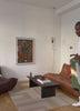 Et lyst og moderne stueområde med minimalistisk indretning, herunder en brun lædersofa, mørkt sofabord, lys gulvtæppe og farverig abstrakt kunst på væggen. Til højre ses en mand delvist i billedet, iført en hvid t-shirt og holdende en farverig genstand.